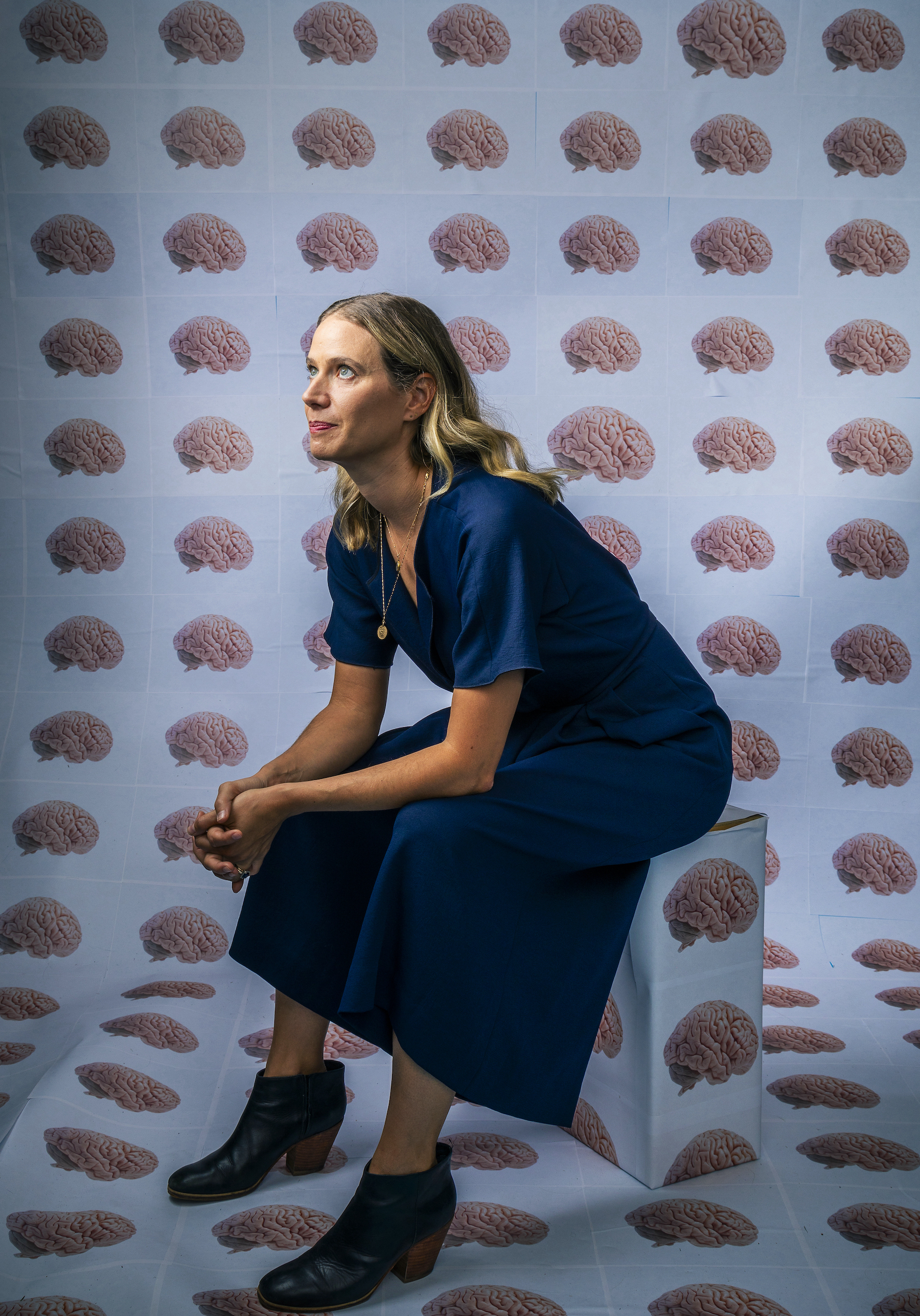 Emily Jacobs sits on a box with hands clasped resting on her knees. She is looking up off to the side of the image. Behind her and covering the box is wall paper that has rows and columns of human brains on a light blue background.
