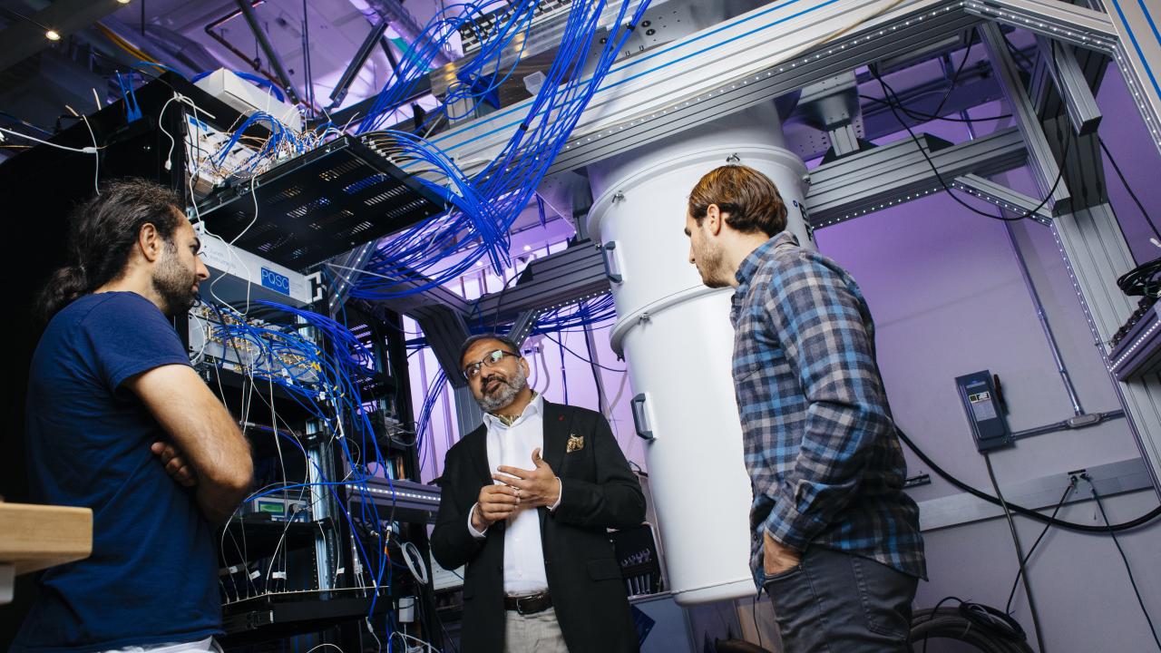 Three men stand talking in a room full of large-scale computers with webs of blue wires daisy chained overhead to various towers.