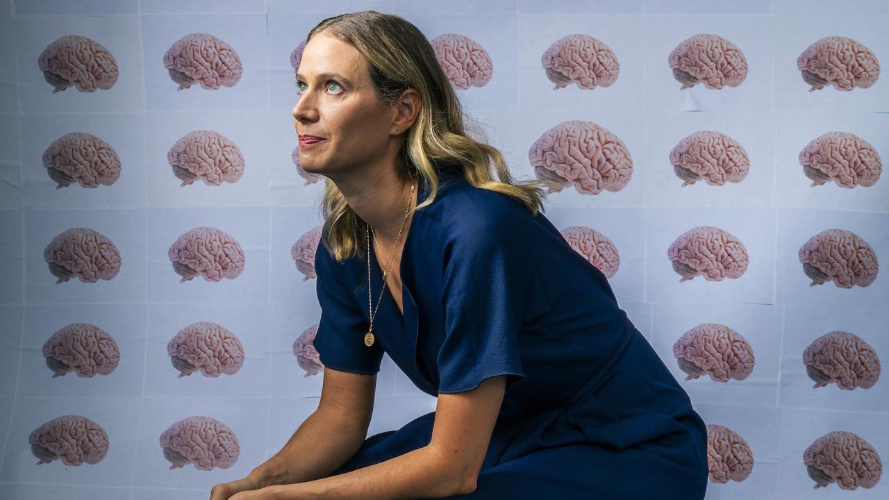Emily Jacobs sits on a box with hands clasped resting on her knees. She is looking up off to the side of the image. Behind her and covering the box is wall paper that has rows and columns of human brains on a light blue background.