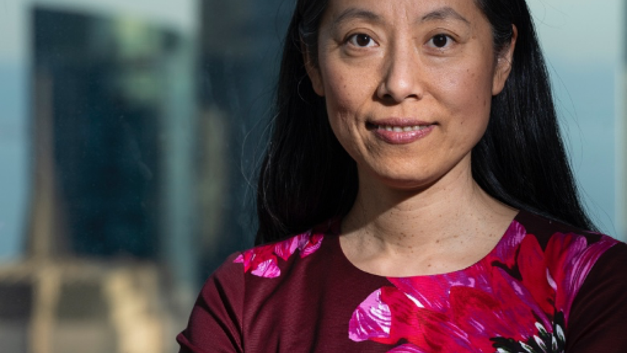 Asian woman with long black hair wearing a dress with large, brightly colored pink flowers printed on it, stands with arms crossed front of a cityscape.