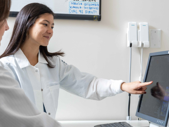 Two women wearing white lab coats look at a computer screen. One woman with long hair is pointing at the screen.