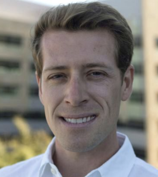 Portrait of Michael Keiser, Ph.D wearing a white collared shirt with tall, tan building behind him in the background.