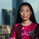 Asian woman with long black hair wearing a dress with large, brightly colored pink flowers printed on it, stands with arms crossed front of a cityscape.